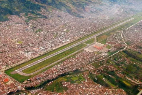 Wide aerial view showing the long runway of Cusco airport surrounded by the dense urban cityscape and green mountains.