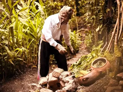 A man in traditional Andean clothing and a chullo hat preparing earth-baked food in clay pots next to a stone oven