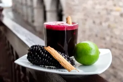 A glass of Chicha Morada served with purple corn, cinnamon sticks, and lime.