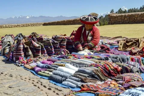 An indigenous woman in traditional Andean clothing displaying colorful handmade textiles at the Chinchero market with snow-capped mountains in the background.