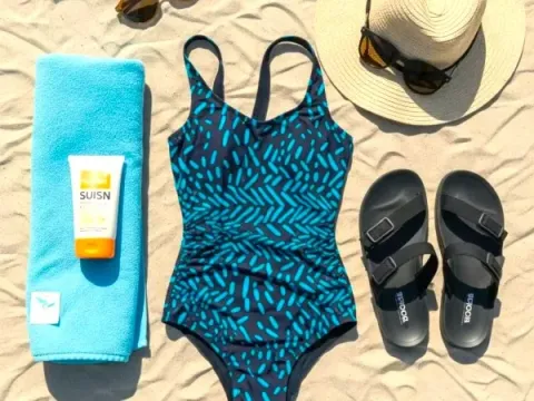 Blue one-piece swimsuit with leaf print, sandals, towel, and hat, on the sand at the beach