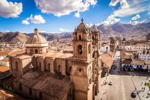 Beautiful aerial view of the Cusco Cathedral and colonial red-tiled roofs under a blue sky with the Andes mountains in the background.