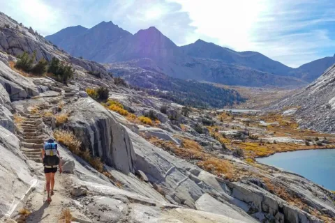 A lone hiker walking down stone steps on a mountain trail overlooking a blue lake on the John Muir Trail.