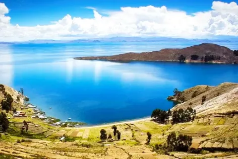 A wide scenic view of the blue Lake Titicaca with yellow terraced hills in the foreground under a bright cloudy sky.
