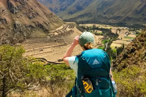 A traveler with a teal backpack overlooks the ancient Inca ruins of Llactapata in the valley below during a sunny day on the trail.