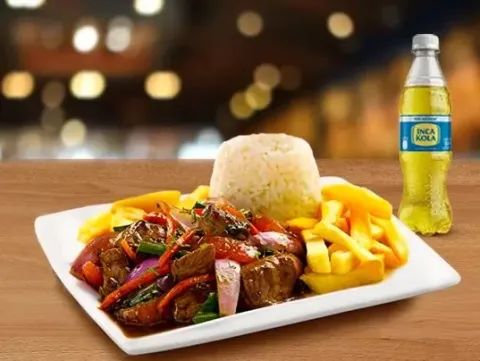 A plate of Lomo Saltado with rice and french fries, accompanied by a bottle of Inca Kola soda.