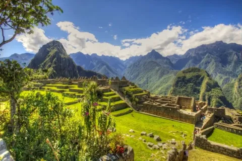 Wide panoramic view of Machu Picchu ruins, green terraces, and the surrounding mountain peaks under a blue sky.