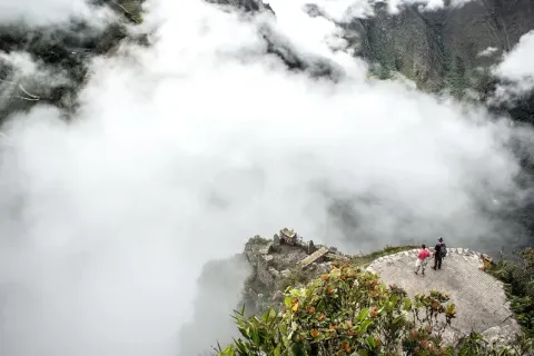 Two travelers standing on a stone lookout point surrounded by heavy fog, with the faint outline of Inca ruins appearing through the clouds.