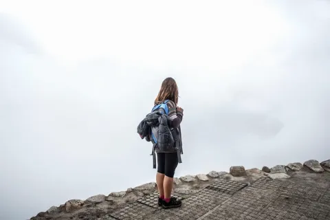 A female hiker with a backpack stands on a stone path looking at the dense white fog covering the mountains of Machu Picchu.