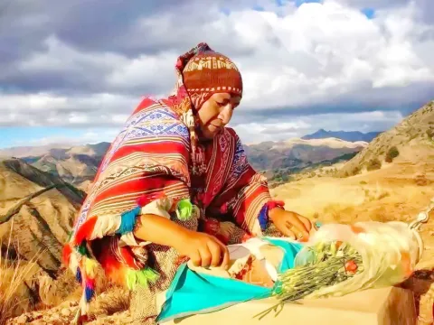 An Andean woman in a colorful poncho performing a ritual offering to Mother Earth (Pachamama) with a mountain landscape in the background