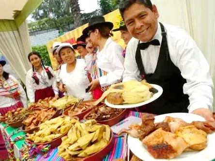 A smiling waiter in a formal vest serving plates of Pachamanca meat and tamales at a colorful outdoor event