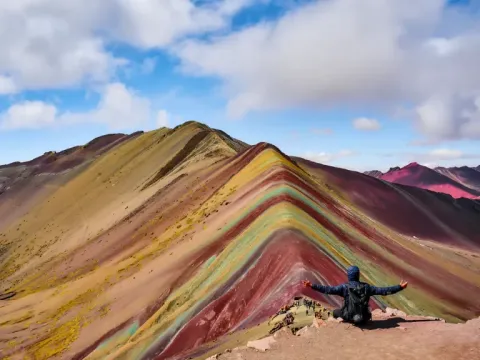 Person sitting with arms open at the top of Vinicunca Mountain, capturing a panoramic view