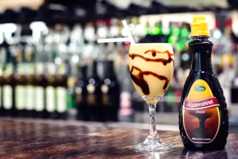 A glass of Algarrobina cocktail next to a bottle of sweet carob syrup on a bar.