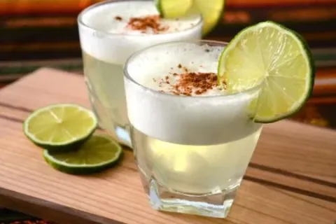 Two glasses of Pisco Sour cocktail topped with foam, cinnamon, and a lime slice on a wooden board.