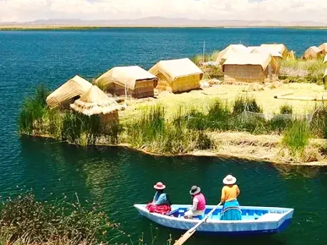 Three people in a blue rowing boat near a small floating house made of totora reeds in the Uros Islands.