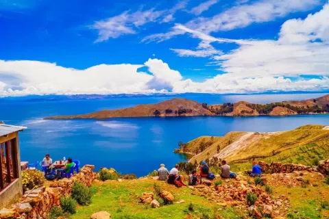 A group of travelers sitting on a stone wall at a viewpoint overlooking the deep blue Lake Titicaca and distant mountains.
