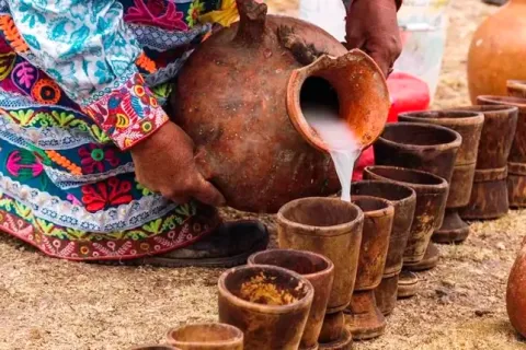 An indigenous woman pouring Chicha de Jora from a clay pitcher into wooden cups.