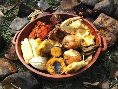 A clay bowl filled with traditional Peruvian Pachamanca including corn, potatoes, fava beans, and various meats cooked underground