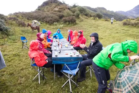 A group of trekkers in colorful rain gear enjoying an outdoor lunch at a long table during a mountain expedition.