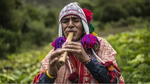 Man in traditional dress playing a wind instrument in a natural setting 