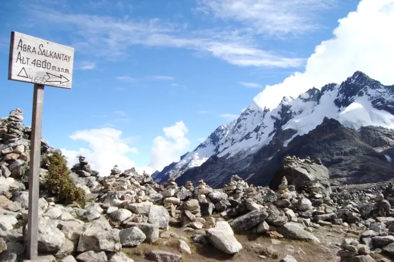 A wooden sign marking Abra Salkantay at 4,600 meters altitude surrounded by stone cairns and snowy peaks.