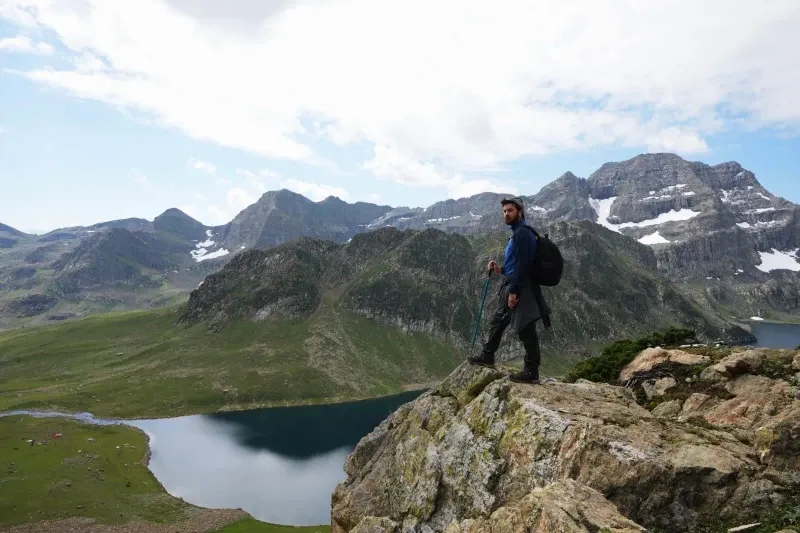 A hiker with a backpack standing on a rocky outcrop overlooking a deep blue mountain lagoon surrounded by green peaks.