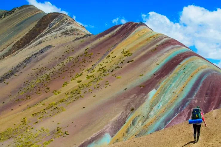 A hiker with a backpack and blue sleeping mat walking up the trail toward the colorful slopes of Vinicunca.
