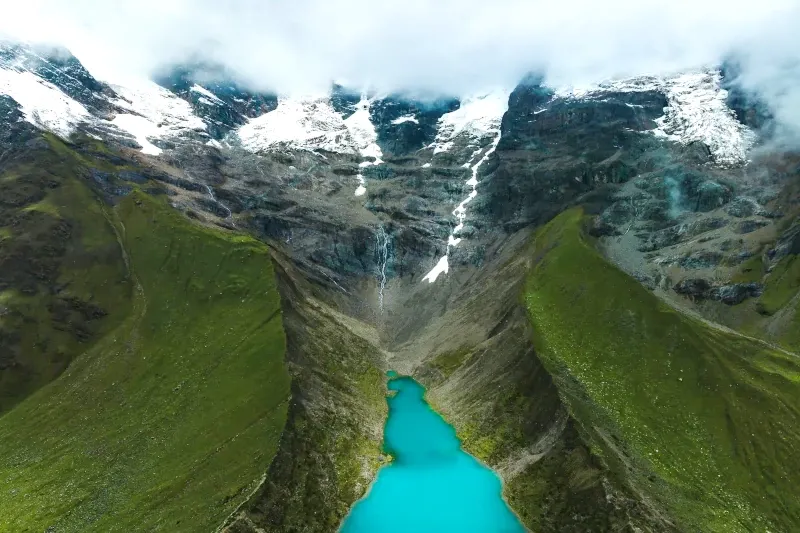 An aerial perspective of the vibrant turquoise Humantay Lake surrounded by steep green mountain ridges and glaciers.