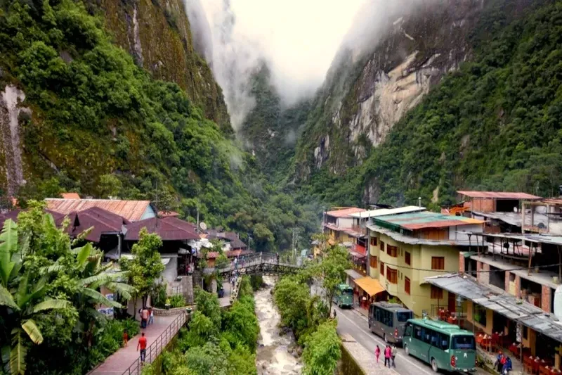 Buses parked on the main road of Aguas Calientes with misty green mountains in the background.