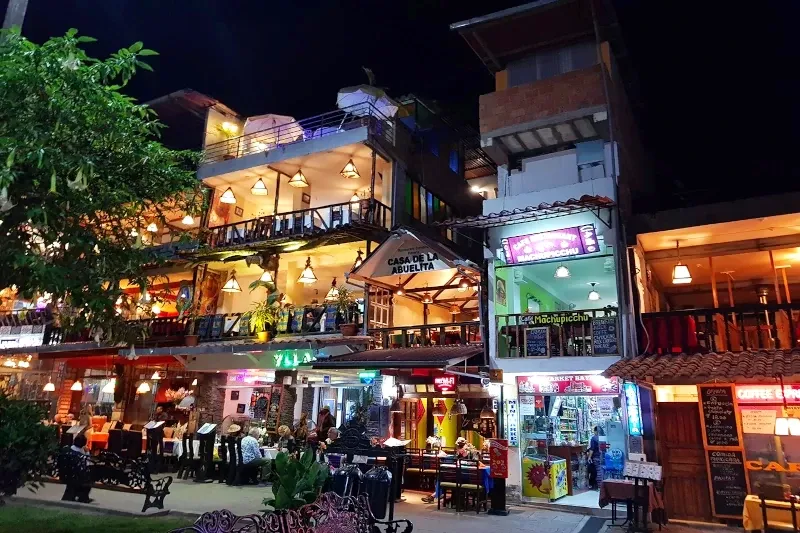 Brightly lit multi-story restaurants and shops along a busy street in Aguas Calientes at night.