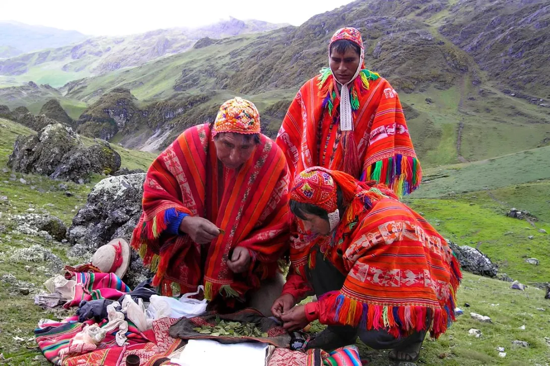 Three Quechua men in traditional red ponchos performing a sacred coca leaf ceremony in the Andean highlands.