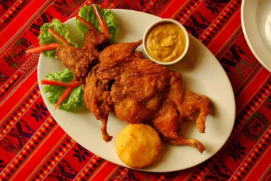 High-angle shot of a traditional Cuy Chactado dish on an Andean textile, featuring a whole fried guinea pig with lettuce garnishing and yellow chili sauce.