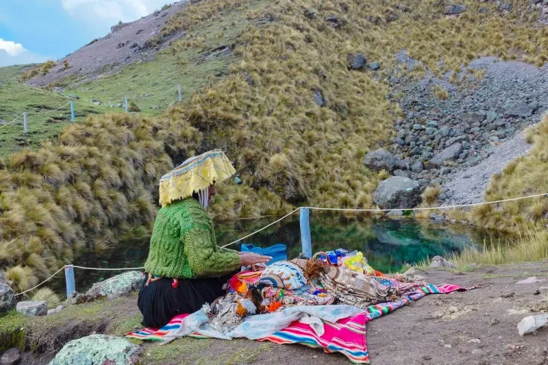 A local Andean woman in traditional green clothing and a decorated hat selling colorful handmade textiles by a turquoise mountain lake.