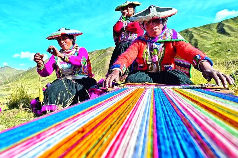 Indigenous women in traditional Montera hats using a traditional loom to create colorful Peruvian textiles in the highlands.