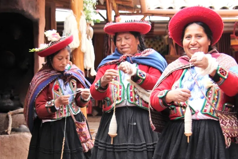 Three smiling Andean women in traditional colorful dress spinning wool using a pushka in a workshop in Cusco.
