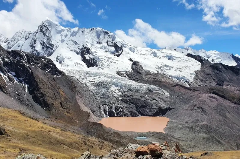 A high-altitude red lagoon situated at the base of a massive glacier on the Ausangate mountain range under a blue sky.