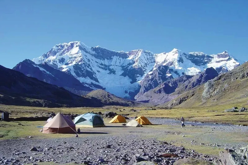 Colorful camping tents set up in a valley facing a massive glacier-covered mountain range under a clear sky.