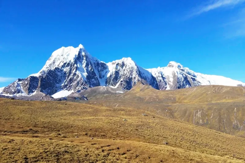 The massive snow-capped Ausangate mountain rising above the dry Andean highlands under a bright blue sky.