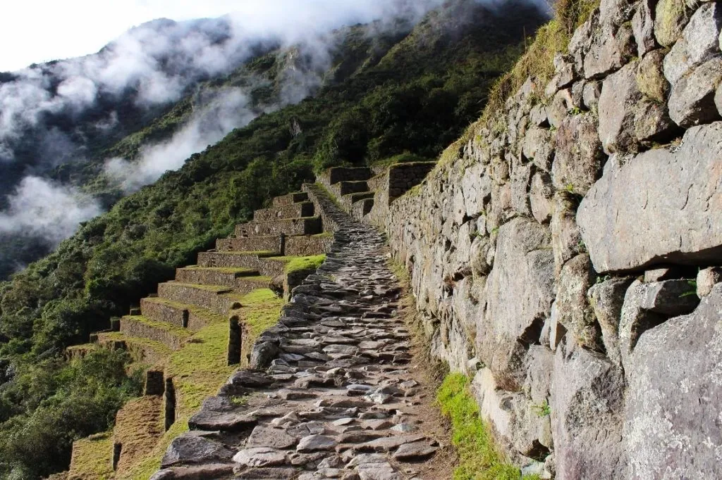 A stone-paved path of the Inca Trail next to agricultural terraces with clouds in the valley.