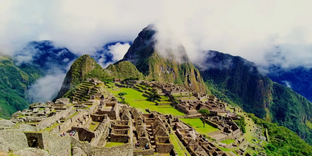 Iconic panoramic view of the Machu Picchu citadel surrounded by misty mountains and clouds under a bright sky.