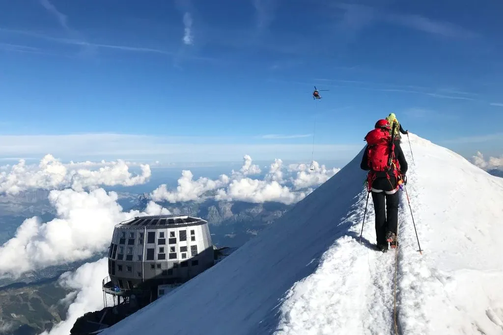 A mountaineer with a red backpack walking along a narrow snowy ridge towards a mountain hut at Mont Blanc.