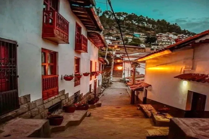 A narrow colonial street in Cusco at dusk with warm street lights, white walls, and red wooden balconies.