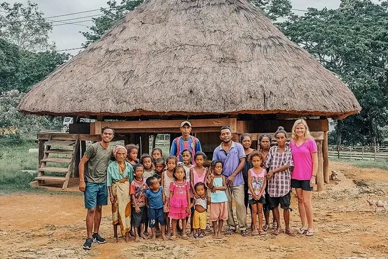 Group of tourists posing with a local indigenous family and children in front of a traditional thatched-roof jungle hut.