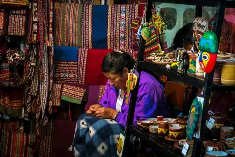 Authentic handmade crafts and artisans in Cusco. A local artisan in a purple jacket carefully hand-sewing traditional crafts surrounded by colorful textiles and Peruvian masks.