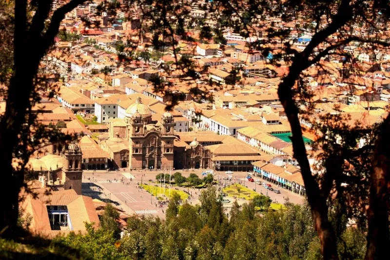 A high-angle view framed by trees showing the Cusco Cathedral and the historic Plaza de Armas surrounded by colonial buildings.