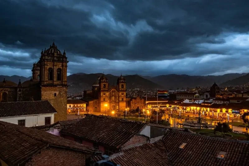 A night view of the Plaza de Armas in Cusco, showing the illuminated Cathedral and colonial architecture under a dramatic dark sky.