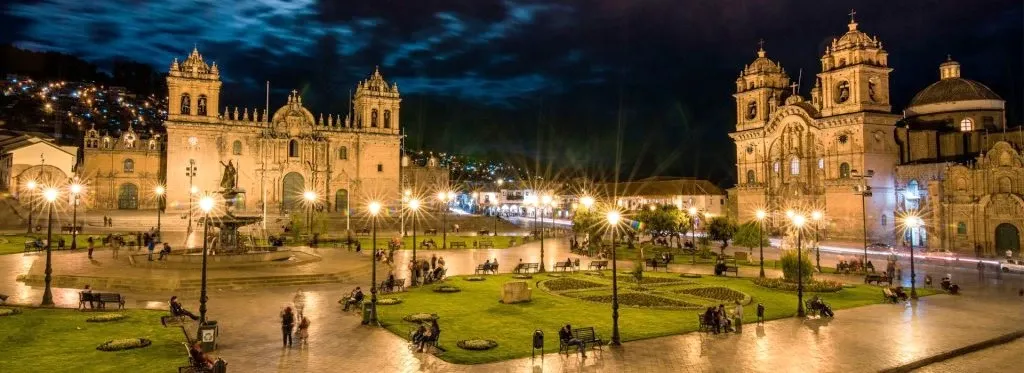 Stunning night view of Plaza de Armas in Cusco with the illuminated Cathedral and Church of the Society of Jesus under a dramatic evening sky.
