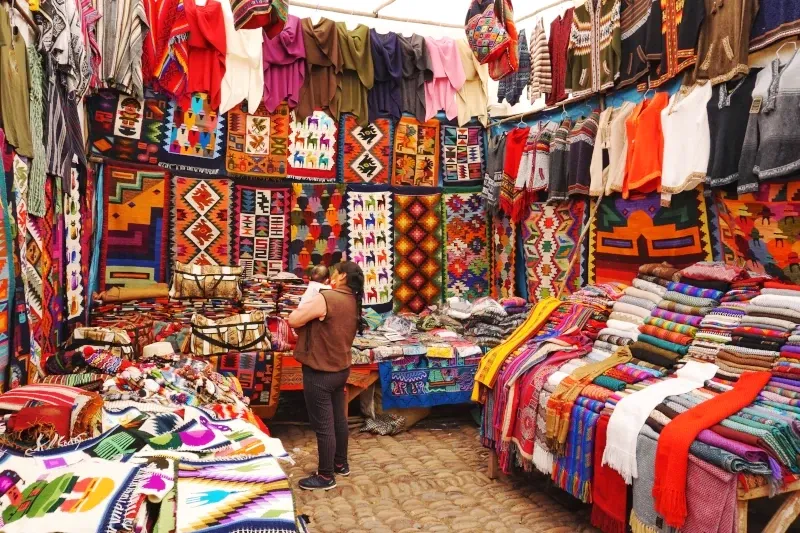 A local woman carrying a baby while standing in a colorful market stall filled with traditional Andean textiles and ponchos.