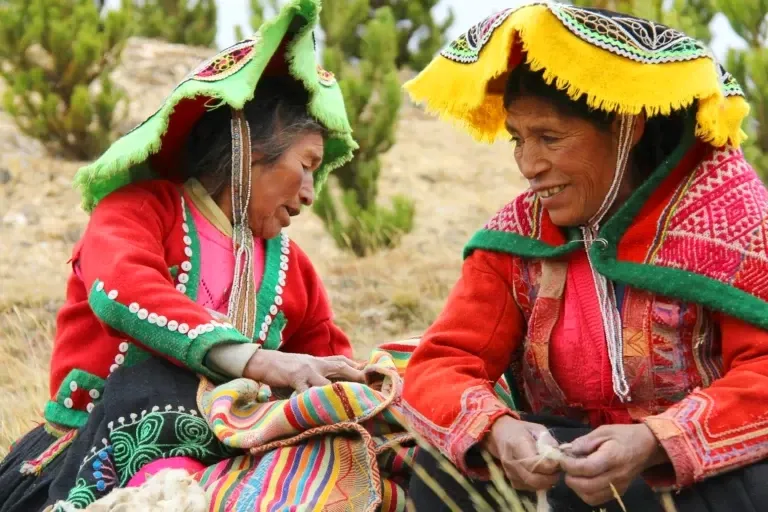 Two indigenous Andean women in vibrant red and green traditional clothing weaving colorful textiles outdoors.