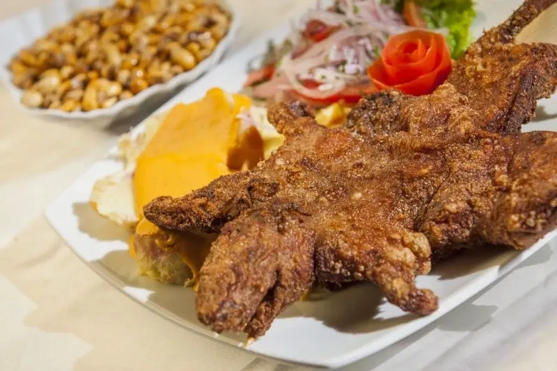 A close-up of fried guinea pig served with Huancaina creamy sauce, toasted corn (cancha), and a decorative tomato rose salad.
